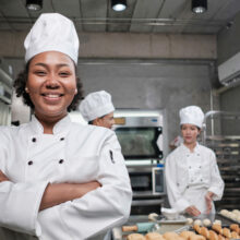 Young African American female chef in white cooking uniform looks at camera, arms crossed and cheerful smile with food professional occupation, commercial pastry culinary jobs in a restaurant kitchen.